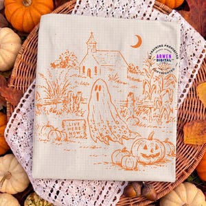 May include: A cream-colored fabric square featuring an orange line-art illustration of a ghost, a church, pumpkins, and a sign that reads "LIVE HERE NOW". The design includes a crescent moon and corn stalks, all set against a subtle grid pattern.