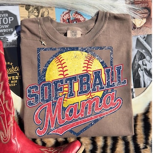 May include: A brown Comfort Colors t-shirt featuring a softball design. The design includes a yellow softball with red stitching, and the words "SOFTBALL Mama" in blue and red lettering. The shirt is displayed on a textured surface.