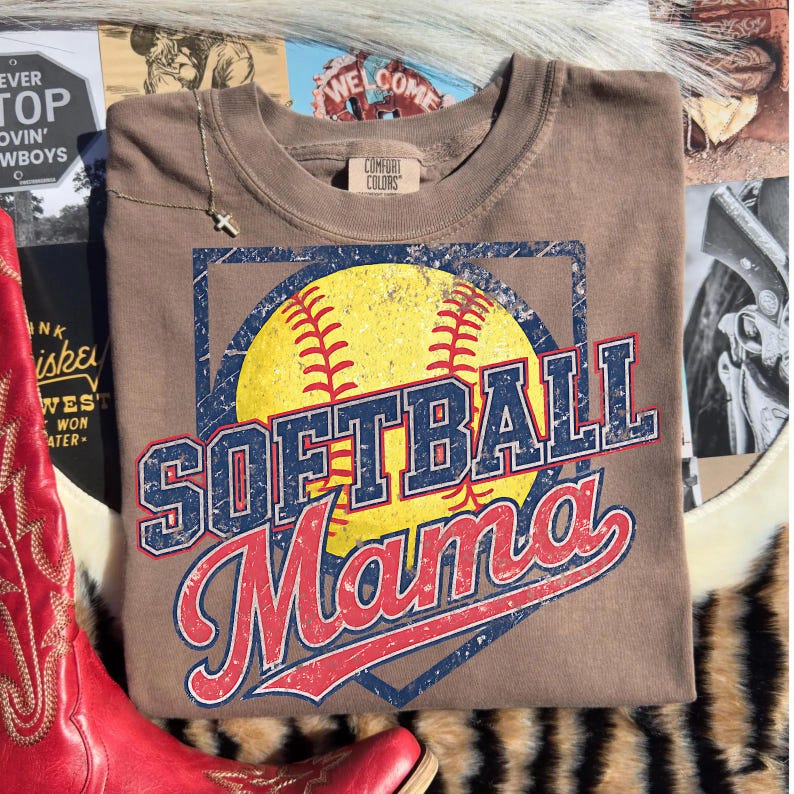 May include: A brown Comfort Colors t-shirt featuring a softball design. The design includes a yellow softball with red stitching, and the words "SOFTBALL Mama" in blue and red lettering. The shirt is displayed on a textured surface.
