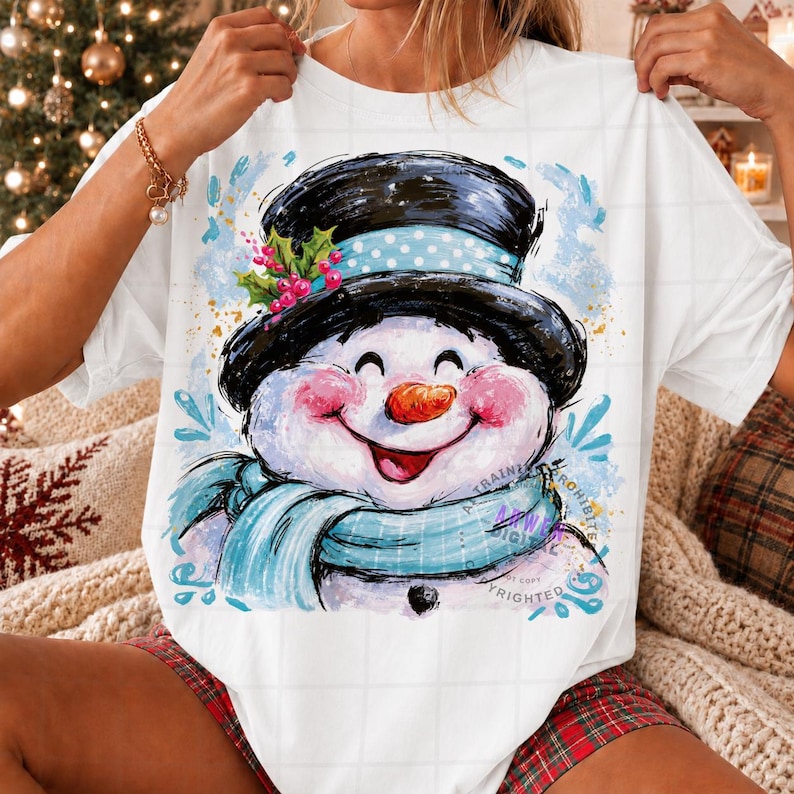 May include: A white t-shirt featuring a graphic illustration of a smiling snowman wearing a black top hat with a light blue polka dot band, green holly leaves, and red berries. The snowman has a bright orange carrot nose and a light blue scarf.
