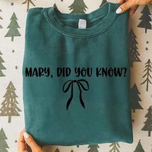 Może przedstawiać: Turkusowa bluza z napisem "MARY, DID YOU KNOW?" w kolorze czarnym, wraz z czarną grafiką kokardy. Bluza jest trzymana w dłoniach.