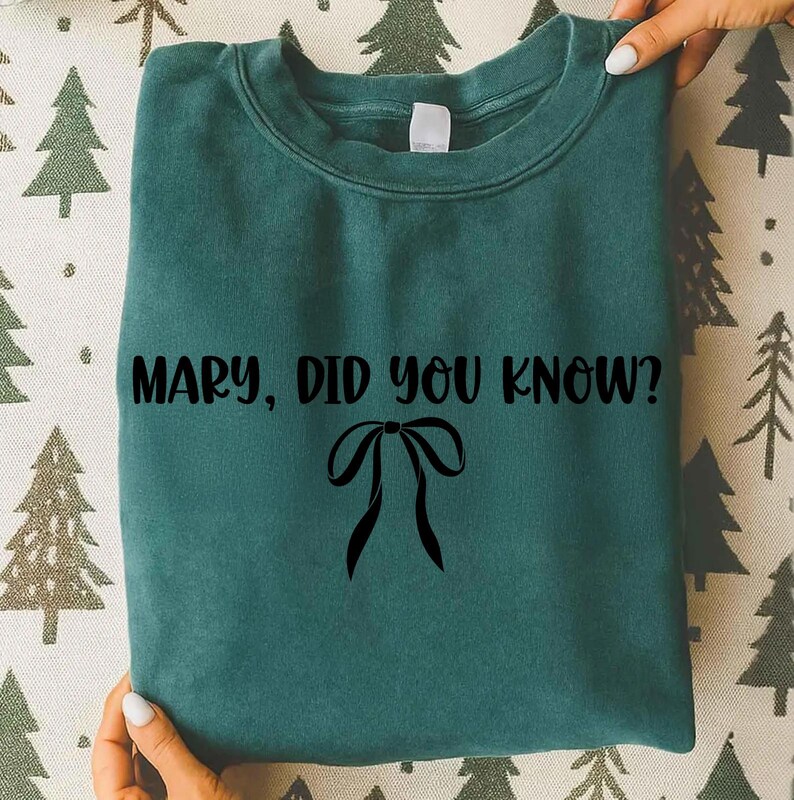 Może przedstawiać: Turkusowa bluza z napisem "MARY, DID YOU KNOW?" w kolorze czarnym, wraz z czarną grafiką kokardy. Bluza jest trzymana w dłoniach.