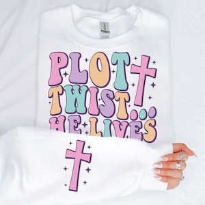 May include: White sweatshirt with a pastel colored graphic that says "Plot Twist... He Lives" with a pink cross underneath.