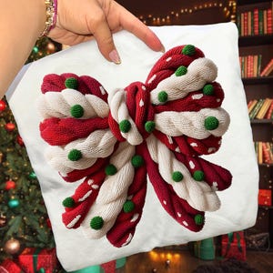May include: White square pillow with a large, beaded bow design. The bow features red and white twisted sections with white polka dots and green beaded accents. The pillow is held by a hand.