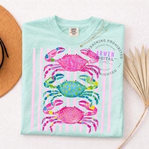 Puede incluir: Camiseta azul claro con un dise&ntilde;o de acuarela de tres cangrejos coloridos apilados verticalmente. Los cangrejos son rosas, azules y rosas, con rayas verticales rosas. La camiseta tiene una etiqueta Comfort Colors.