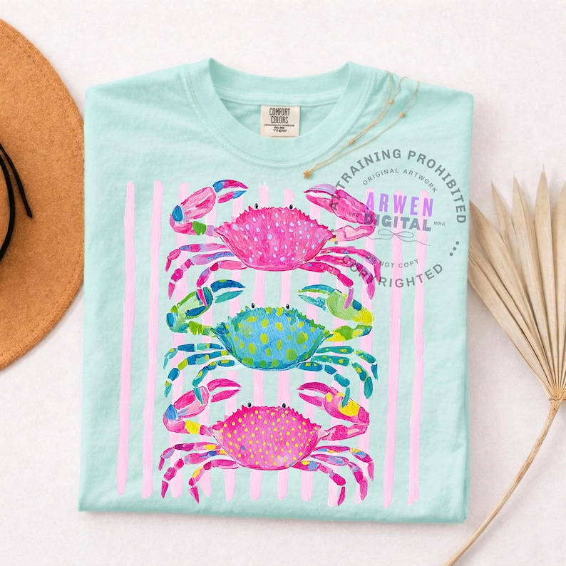 Puede incluir: Camiseta azul claro con un dise&ntilde;o de acuarela de tres cangrejos coloridos apilados verticalmente. Los cangrejos son rosas, azules y rosas, con rayas verticales rosas. La camiseta tiene una etiqueta Comfort Colors.