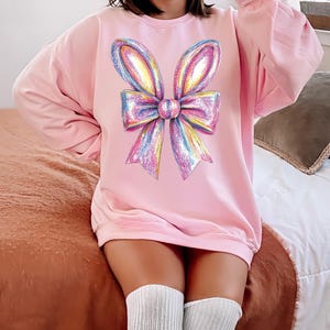 May include: A pink oversized sweatshirt with a large rainbow glitter bow graphic on the front.