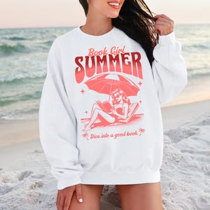 May include: White sweatshirt with a pink graphic design of a woman reading a book on a beach under an umbrella. The text on the sweatshirt reads "Book Girl Summer" and "Dive into a good book."