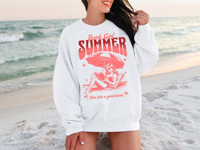 May include: White sweatshirt with a pink graphic design of a woman reading a book on a beach under an umbrella. The text on the sweatshirt reads "Book Girl Summer" and "Dive into a good book."