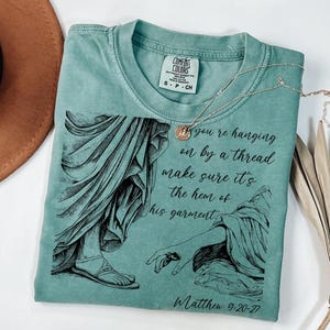 May include: A teal blue t-shirt with a black and white illustration of a hand reaching out to touch the hem of a garment. The text "If you're hanging on by a thread make sure it's the hem of his garment" and "Matthew 9:20-27" are printed on the shirt.