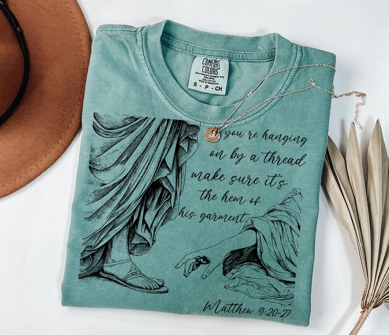 May include: A teal blue t-shirt with a black and white illustration of a hand reaching out to touch the hem of a garment. The text "If you're hanging on by a thread make sure it's the hem of his garment" and "Matthew 9:20-27" are printed on the shirt.
