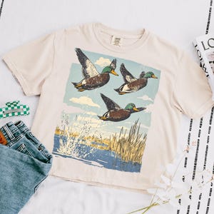 May include: Cream-colored t-shirt featuring a graphic of three ducks in flight over a body of water with reeds. The shirt is paired with blue jeans and a green and white checkered hair clip. A magazine and flowers are also in the image.