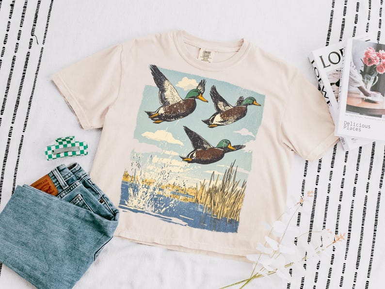 May include: Cream-colored t-shirt featuring a graphic of three ducks in flight over a body of water with reeds. The shirt is paired with blue jeans and a green and white checkered hair clip. A magazine and flowers are also in the image.