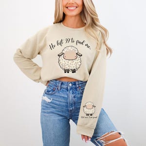 K&ouml;nnte beinhalten: Ein beiger, kurz geschnittener Pullover mit dem Text "He left 99 to find me." und einem Cartoon-Bild eines wei&szlig;en Schafs mit schwarzen Flecken. Der Pullover hat lange &Auml;rmel und einen Rundhalsausschnitt.