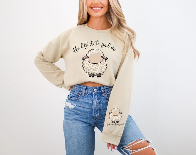 K&ouml;nnte beinhalten: Ein beiger, kurz geschnittener Pullover mit dem Text "He left 99 to find me." und einem Cartoon-Bild eines wei&szlig;en Schafs mit schwarzen Flecken. Der Pullover hat lange &Auml;rmel und einen Rundhalsausschnitt.