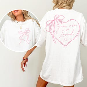 May include: White oversized t-shirt with a pink ribbon bow design on the front and a pink ribbon heart design on the back with the text "you are so loved John 3:16".