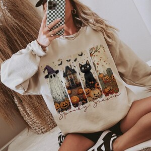 May include: Beige sweatshirt with a Halloween-themed graphic. The graphic includes a ghost, haunted house, black cat, pumpkins, and the words "Spooky Season". A white baseball cap with "Malibu Cowboy" is also visible.