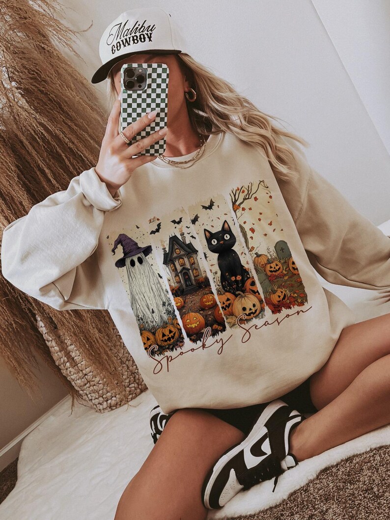 May include: Beige sweatshirt with a Halloween-themed graphic. The graphic includes a ghost, haunted house, black cat, pumpkins, and the words "Spooky Season". A white baseball cap with "Malibu Cowboy" is also visible.