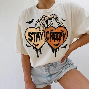 May include: Cream-colored t-shirt featuring a Halloween-themed design. The graphic includes two heart-shaped designs with the words "STAY CREEPY" and a skeleton hand holding the hearts. Bats and dripping details complete the spooky look.