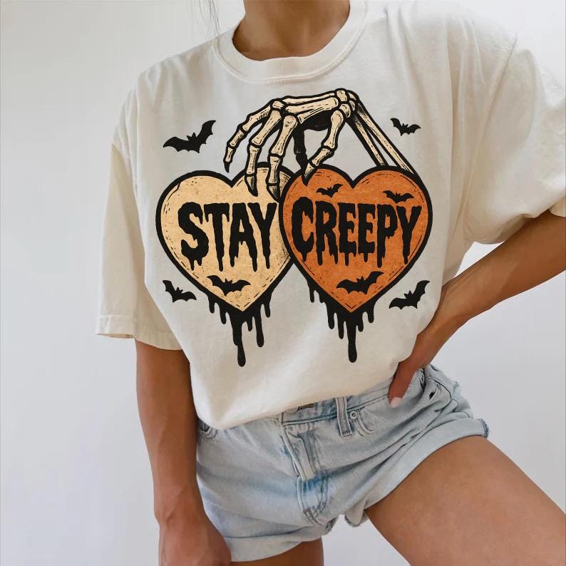 May include: Cream-colored t-shirt featuring a Halloween-themed design. The graphic includes two heart-shaped designs with the words "STAY CREEPY" and a skeleton hand holding the hearts. Bats and dripping details complete the spooky look.