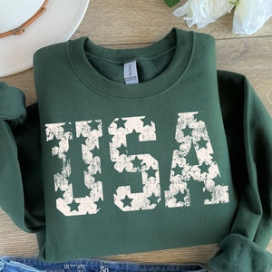 K&ouml;nnte beinhalten: Ein dunkelgr&uuml;ner Pullover mit einem wei&szlig;en, abgenutzten Aufdruck des Wortes "USA", das aus Sternen besteht.