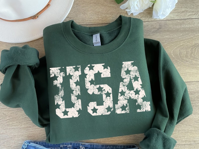K&ouml;nnte beinhalten: Ein dunkelgr&uuml;ner Pullover mit einem wei&szlig;en, abgenutzten Aufdruck des Wortes "USA", das aus Sternen besteht.