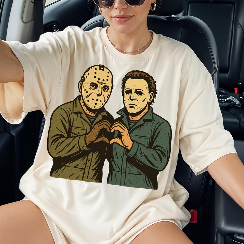 May include: Cream-colored t-shirt featuring a graphic of two figures forming a heart shape with their hands. One figure wears a hockey mask, the other has brown hair. Both are in green jumpsuits.