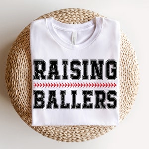May include: White t-shirt with black text that reads "Raising Ballers" with a red baseball stitch design.