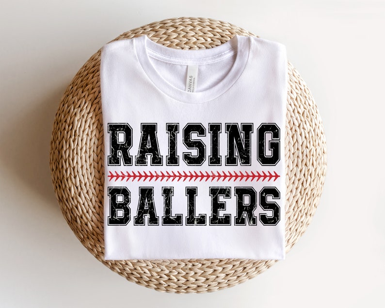 May include: White t-shirt with black text that reads "Raising Ballers" with a red baseball stitch design.