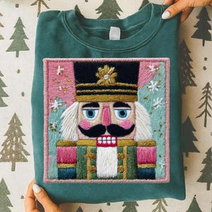 Könnte beinhalten: Ein teal-grünes Sweatshirt mit einem gestickten Nussknacker-Design. Der Nussknacker hat einen schwarzen Hut mit einem goldenen Stern, blaue Augen, einen rosa Schnurrbart und einen roten Mund. Das gestickte Bild ist rosa umrahmt.