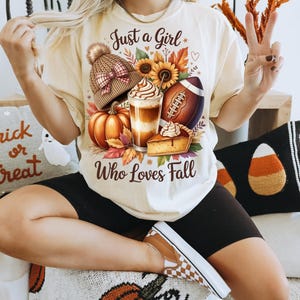 K&ouml;nnte beinhalten: Cremefarbenes T-Shirt mit dem Text "Just a Girl Who Loves Fall". Die Grafik zeigt einen K&uuml;rbis, einen Football, einen Latte, ein St&uuml;ck Kuchen und eine Strickm&uuml;tze. Das Shirt wird mit schwarzen Shorts und karierten Schuhen getragen.