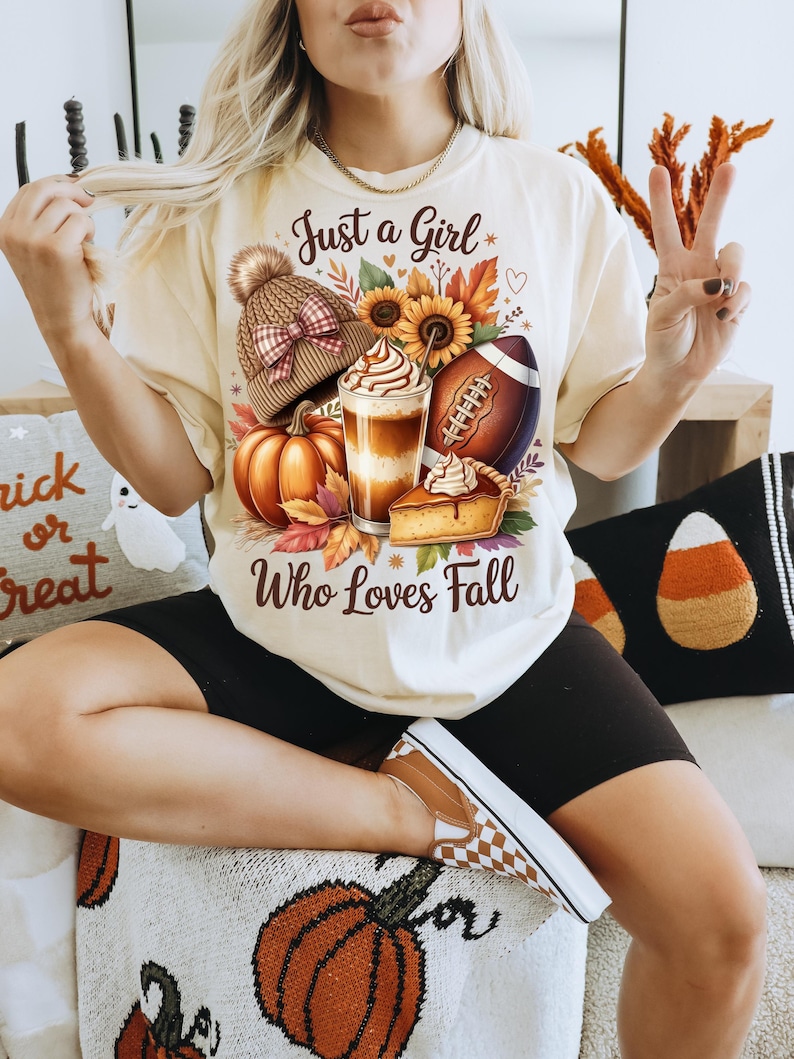 K&ouml;nnte beinhalten: Cremefarbenes T-Shirt mit dem Text "Just a Girl Who Loves Fall". Die Grafik zeigt einen K&uuml;rbis, einen Football, einen Latte, ein St&uuml;ck Kuchen und eine Strickm&uuml;tze. Das Shirt wird mit schwarzen Shorts und karierten Schuhen getragen.