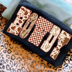 May include: Black sweatshirt with the word "BOOTY" in a western-themed design. The letters feature cow print, cactus, and checkered patterns in brown and cream colors. The sweatshirt is folded on top of other fabric.