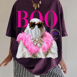 May include: A dark purple t-shirt featuring a graphic of a white ghost wearing leopard print sunglasses and a bright pink feather boa with a large crystal necklace. The word BOO is printed in large, bold, hot pink letters above the ghost.