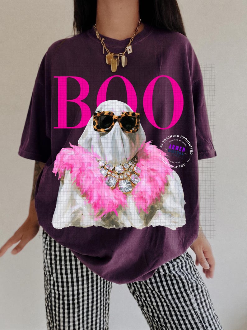 May include: A dark purple t-shirt featuring a graphic of a white ghost wearing leopard print sunglasses and a bright pink feather boa with a large crystal necklace. The word BOO is printed in large, bold, hot pink letters above the ghost.