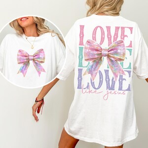 K&ouml;nnte beinhalten: Wei&szlig;es T-Shirt mit einer glitzernden rosa Schleife auf der Vorderseite und dem Text "LOVE LIKE LOVE like jesus" auf der R&uuml;ckseite.