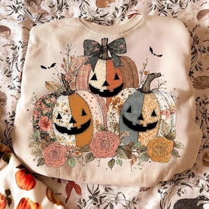May include: Beige sweatshirt featuring a Halloween-themed design. The design includes three smiling pumpkins with floral and plaid patterns, surrounded by flowers and bats. The pumpkins have a patchwork design with a bow on top.