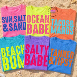 Op de afbeelding: Zes kleurrijke t-shirts met strandthema-zinnen. De shirts zijn roze, geel, oranje, turquoise, paars en blauw. De zinnen zijn onder meer "Sun, Salt & Sand", "Ocean Babe", "Tacos & Margs", "Beach Bun Babe", "Salty Babe" en "Tanned & Tipsy".