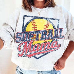 May include: A light gray sweatshirt featuring a distressed graphic of a yellow softball with red stitching, and the words "SOFTBALL Mama" in blue and red lettering. The design is set within a blue square.