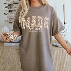Op de afbeelding: Een taupekleurig T-shirt met de tekst "MADE for more" in een bloemenpatroon. De tekst "Ephesians 2:10" staat eronder. Het shirt heeft een losse pasvorm.