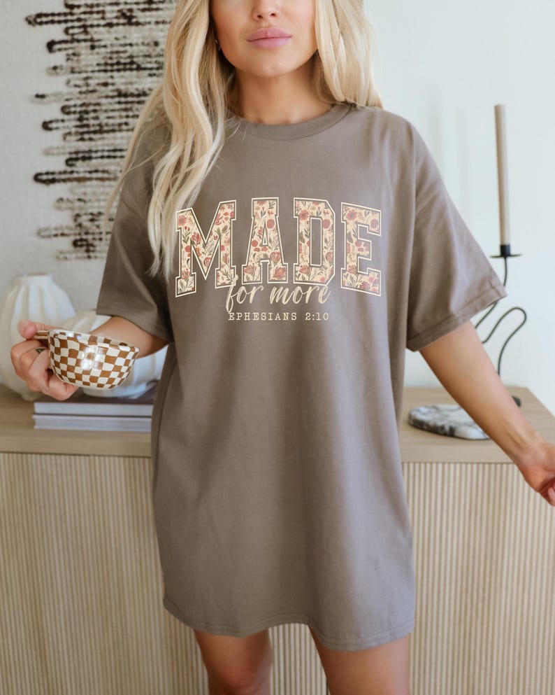 Op de afbeelding: Een taupekleurig T-shirt met de tekst "MADE for more" in een bloemenpatroon. De tekst "Ephesians 2:10" staat eronder. Het shirt heeft een losse pasvorm.