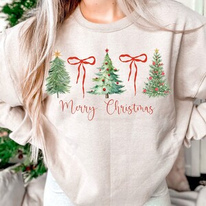 May include: Beige sweatshirt featuring watercolor Christmas trees and red bows. The phrase "Merry Christmas" is written in red script below the trees. A festive holiday-themed top.