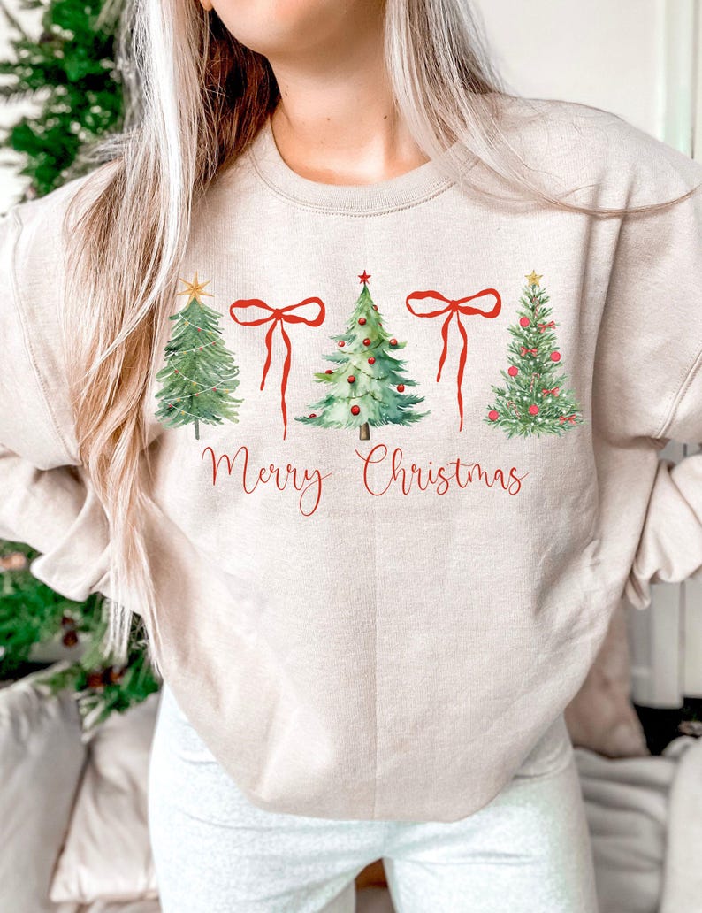 May include: Beige sweatshirt featuring watercolor Christmas trees and red bows. The phrase "Merry Christmas" is written in red script below the trees. A festive holiday-themed top.