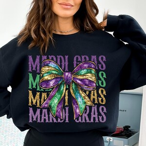 Puede incluir: Sudadera negra con un lazo brillante morado, verde y dorado y el texto "Mardi Gras" repetido tres veces.