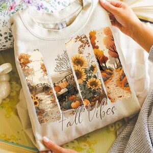 May include: Beige sweatshirt featuring a fall-themed graphic. The design includes sunflowers, pumpkins, and a pathway, with the words "Fall Vibes" in a script font. The sweatshirt is made of a soft material.