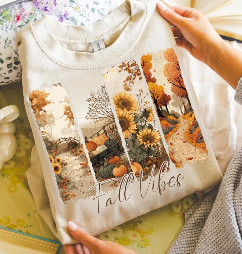 May include: Beige sweatshirt featuring a fall-themed graphic. The design includes sunflowers, pumpkins, and a pathway, with the words "Fall Vibes" in a script font. The sweatshirt is made of a soft material.