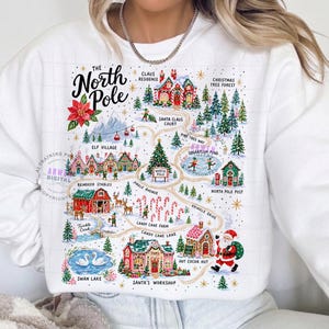 May include: A white long-sleeve sweatshirt featuring a colorful illustrated map of the North Pole. The graphic includes labels for Santa's Workshop, Reindeer Stables, Elf Village, and Candy Cane Lane, with festive imagery of trees, snow, and Santa Claus.