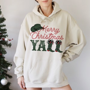May include: A beige hoodie with a festive design. The graphic includes a green cowboy hat, the text "Merry Christmas Y'all" in red and green, and green cowboy boots with candy cane accents. The hoodie has a drawstring hood.