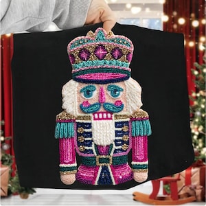 May include: A colorful, embroidered nutcracker design on a black background. The nutcracker features a crown, pink and blue uniform, and a white beard. The design is detailed with gold and teal accents, perfect for holiday decor.