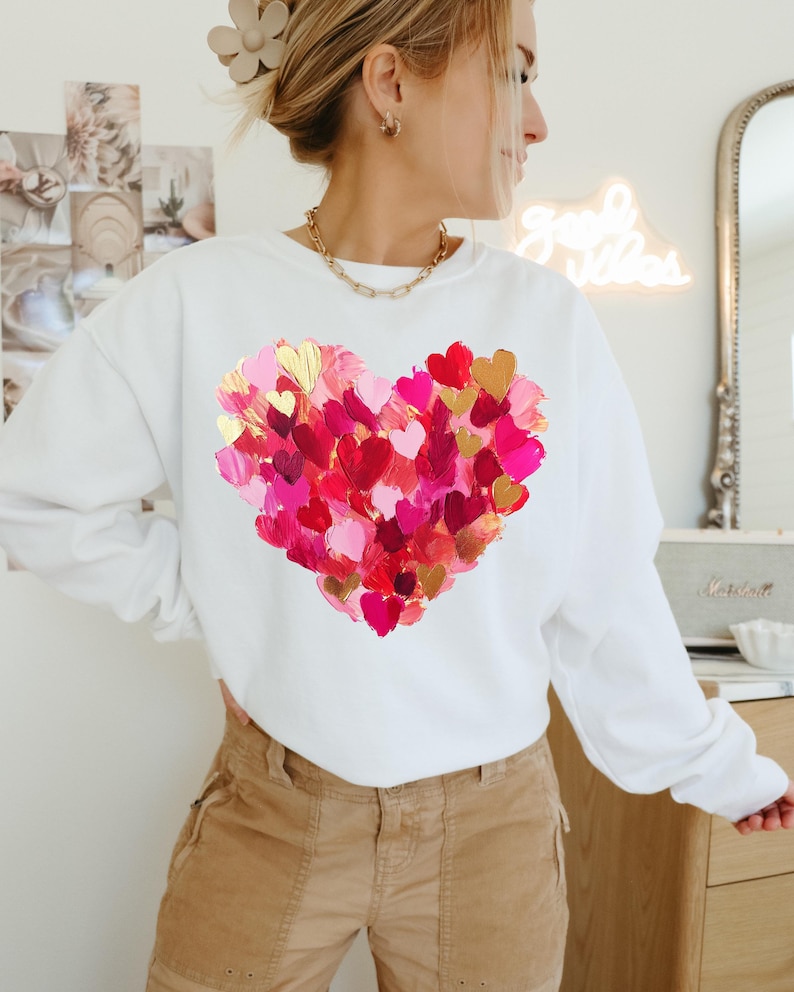 May include: White sweatshirt with a large heart design made of pink, red, and gold painted hearts.